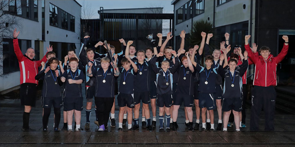 A cheering St John’s Academy Year 9 rugby team, flanked by their supporting Marlborough RFC coaches, proudly show off their medals and the Swindon Schools Rugby Cup